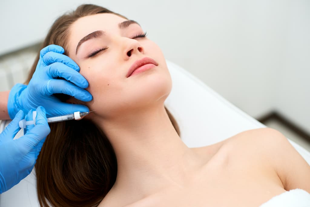Alt text: Woman receiving dermal filler injection in nasolabial fold area by practitioner in blue gloves, demonstrating professional smile line treatment for facial rejuvenation and wrinkle reduction