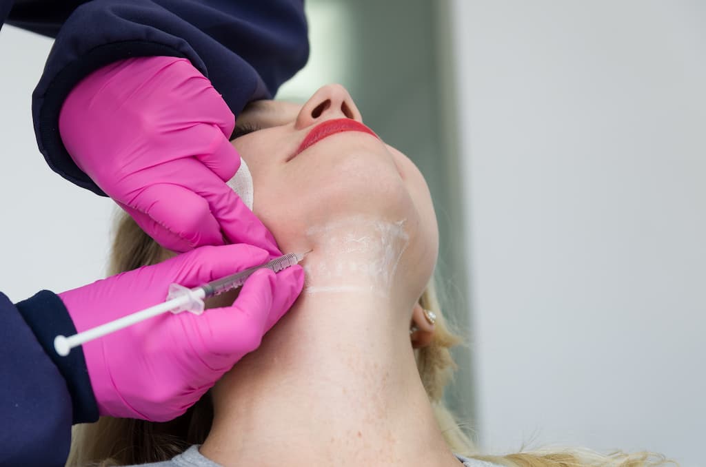 Woman receiving Kybella injection in submental area by practitioner in pink gloves, demonstrating double chin reduction treatment for permanent fat elimination and jawline definition