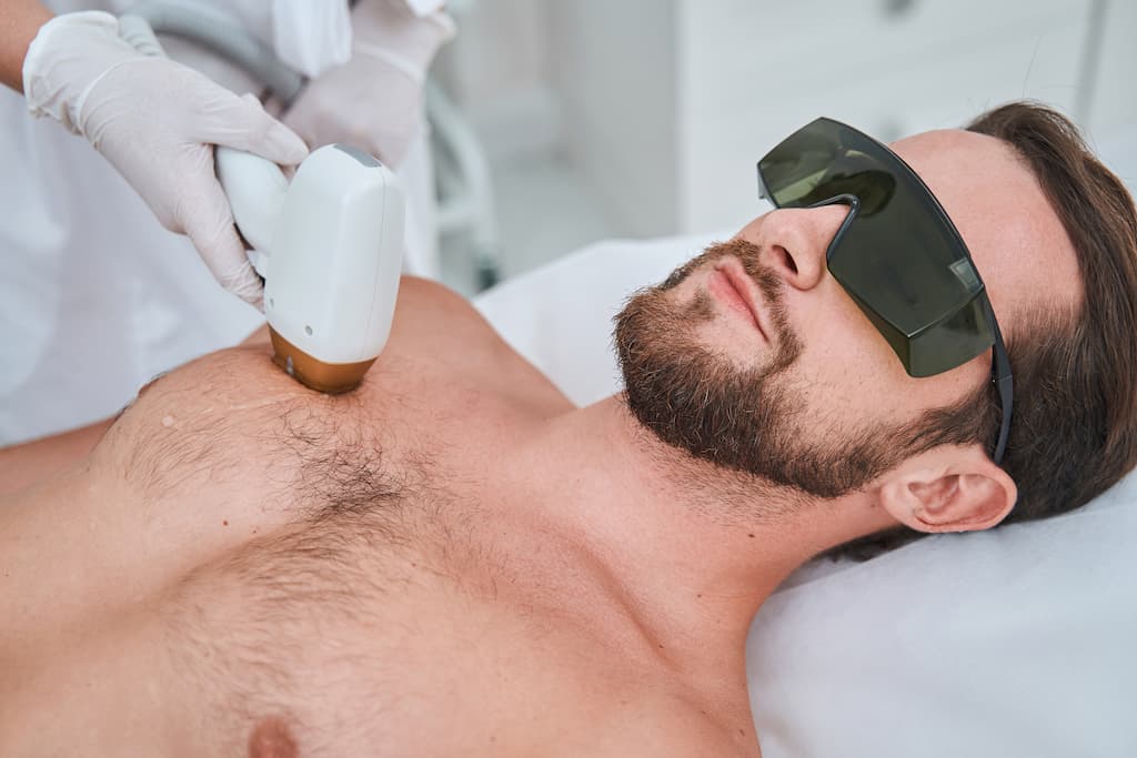 Man receiving laser hair removal treatment on chest area by practitioner in protective eyewear, demonstrating professional male grooming procedure for permanent hair reduction
