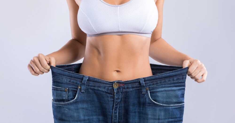 Woman in a white sports bra pulling out the waistband of oversized jeans to show weight loss results