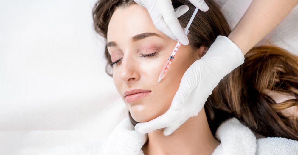 Woman lying down with eyes closed receiving a Botox injection along the jawline from a provider wearing white gloves