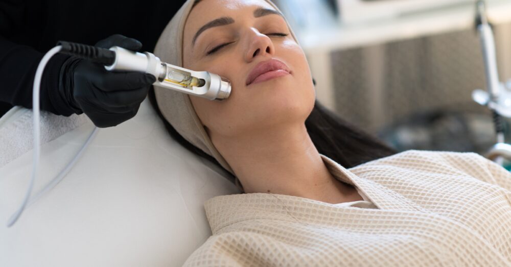 A woman with closed eyes lying back on a treatment bed while a gloved hand holds a HydraFacial wand near her cheek