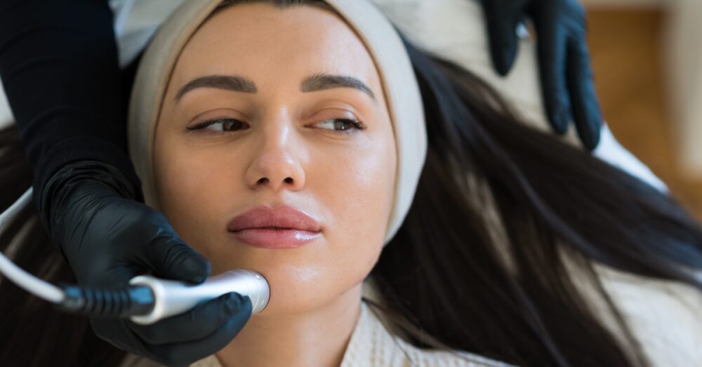 A woman wearing a white headband reclines during a facial treatment as a gloved hand applies a device to her cheek in a clinic setting