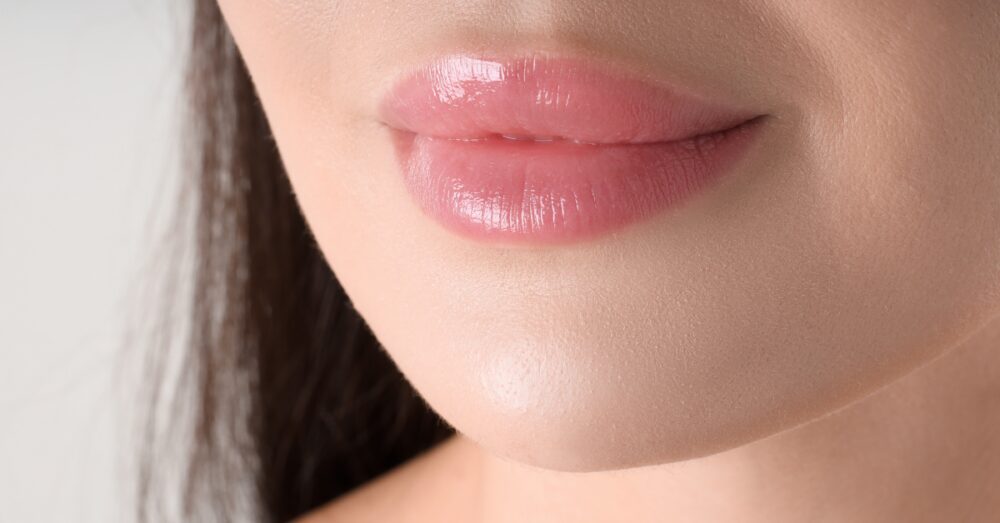 Close up image of a woman’s plump, pink lips, softly smiling against a neutral background