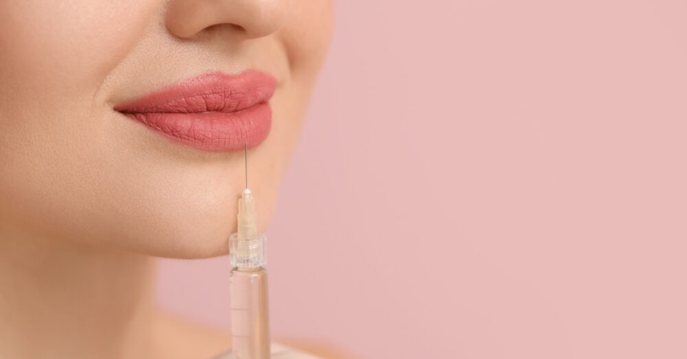 Close up image of a woman receiving lip filler treatment on her lower lip against a pink background